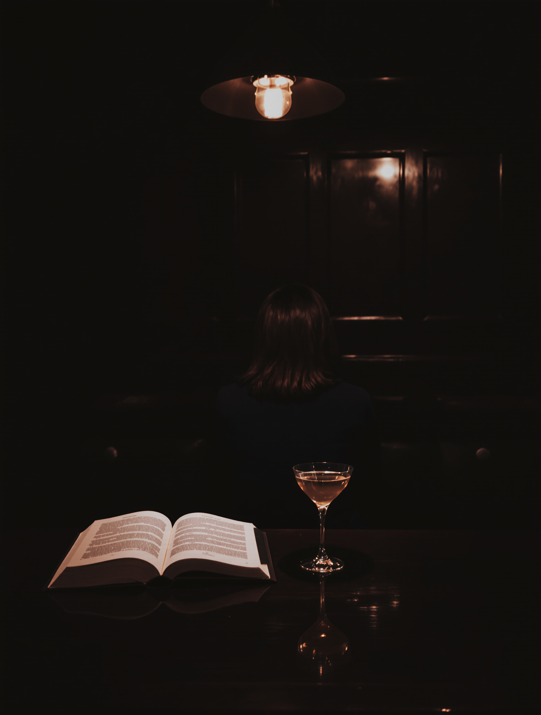 Ambiance feutrée d'un bar à cocktails parisien. Une personne seule, de dos, est assise au comptoir en bois sombre, éclairée par une lampe de style industriel. On devine un livre posé à côté de son verre, suggérant une soirée solo chic et choisie.